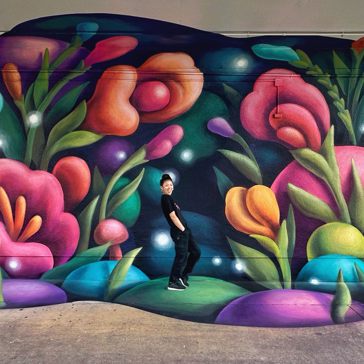 Person standing in front of a colorful mural featuring large, stylized flowers and plants with glowing orbs on a dark background.