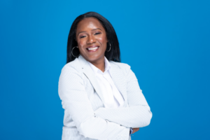 Woman in a white suit jacket and white shirt stands against a blue background, smiling with her arms crossed.