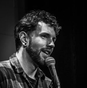 A man with curly hair and a beard speaks into a microphone, wearing a plaid shirt. The image is in black and white.