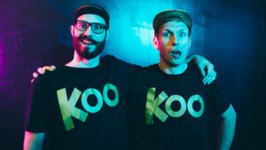 Two men stand side by side, each wearing a black t-shirt with the word "Koo" in gold letters, against a blue and purple-lit background.