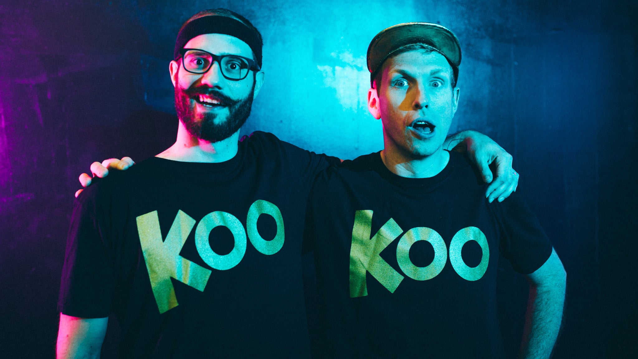 Two men stand side by side, each wearing a black t-shirt with the word "Koo" in gold letters, against a blue and purple-lit background.