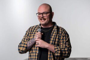 A man with glasses, a mustache, and tattoos holds a microphone and smiles while wearing a plaid shirt and a gold chain, standing against a plain background.