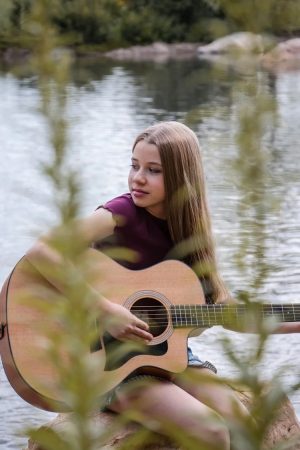 A girl with long hair sits by a lake, holding and playing an acoustic guitar. Green foliage is partially visible in the foreground.