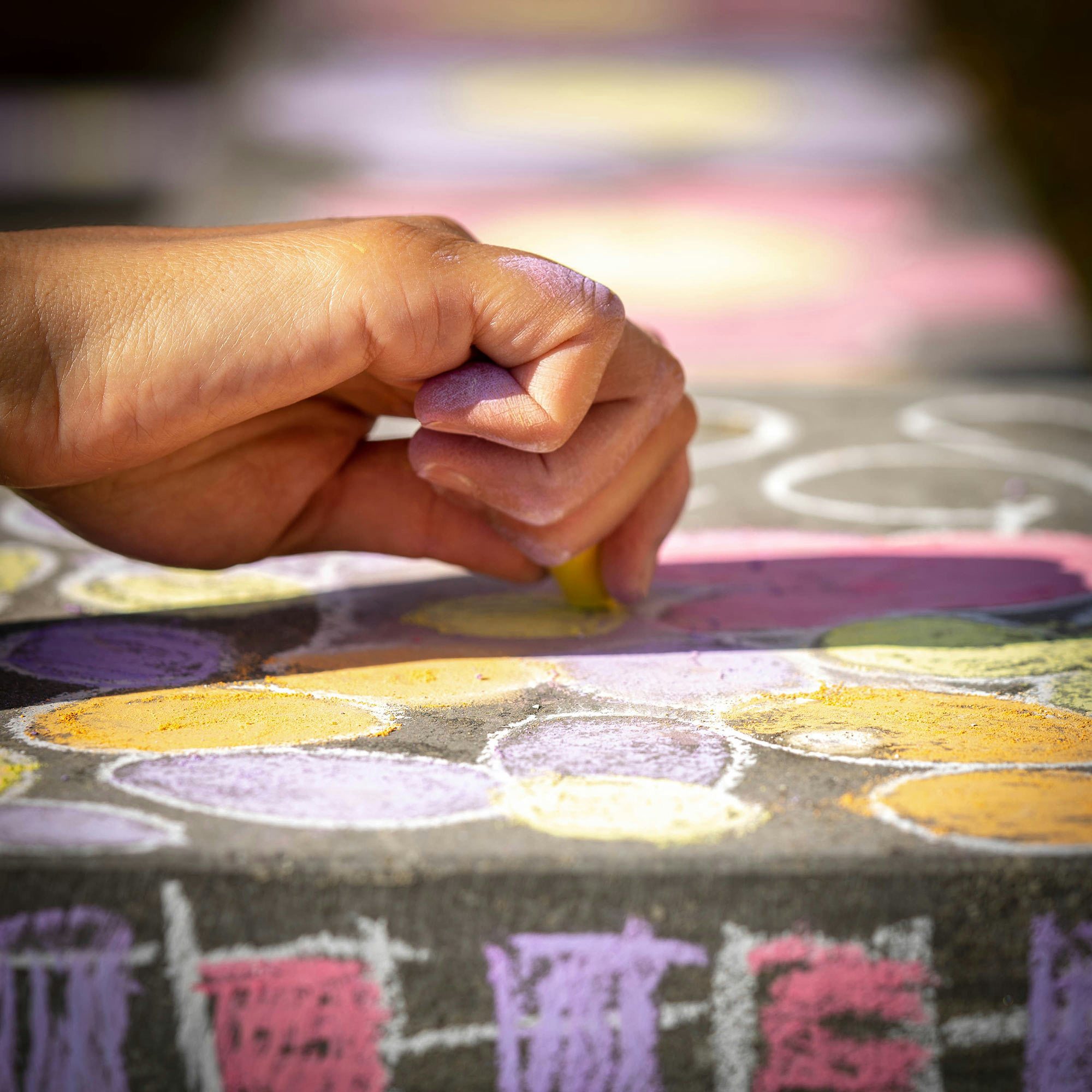 A hand draws colorful circles with chalk on pavement, featuring yellow, purple, and pink shapes outlined in white.