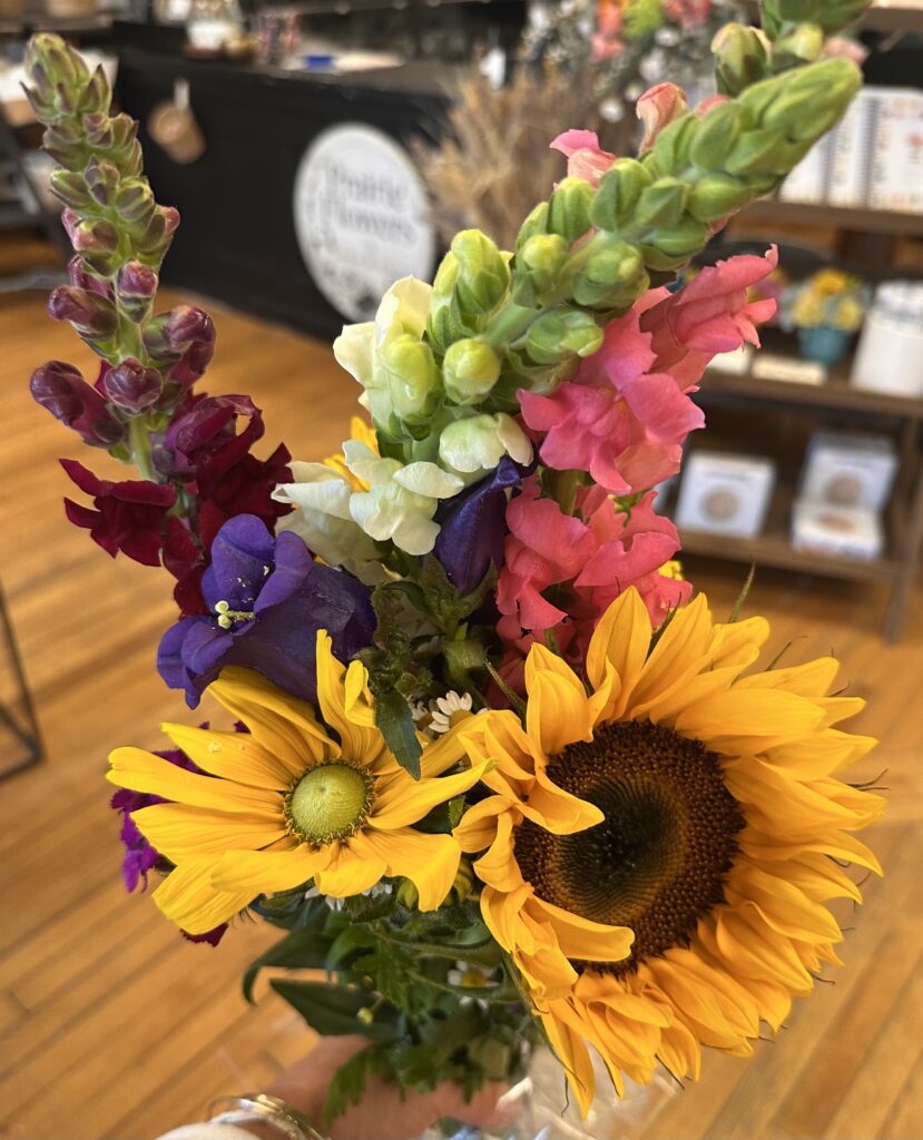 A bouquet featuring a sunflower, yellow daisy, snapdragons in pink, white, and red, and purple flowers, set against a wooden floor and a shop interior background.