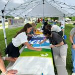 Several people work together painting a large canvas under a white canopy tent at an outdoor mural fest, creating vibrant artwork as part of the community event.