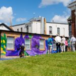 A group of people stands on grass observing a mural of large purple flowers being painted on the side of a small brick building during a sunny mural fest.