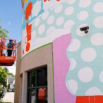 A person on a lift paints a colorful mural with polka dots and abstract shapes on the side of a building during mural fest.