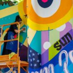 A person on a lift paints a colorful mural with abstract shapes, stars, and the word “sun” on an outdoor wall during a vibrant mural fest.