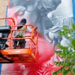 An artist on a lift paints a large mural of a woman’s face and roses in shades of red, white, and gray on a brick building during mural fest.