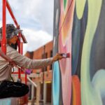 A person on a lift paints a colorful mural on an outdoor wall using a paintbrush, with buildings and a cloudy sky in the background, capturing the vibrant spirit of mural fest.