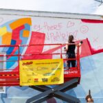 A person on a red lift paints a colorful mural on a building wall during mural fest, with sponsor banners displayed on the lift platform.