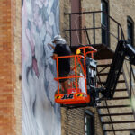 A person in a bucket lift paints a mural on the side of a brick building near metal stairs during mural fest.