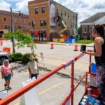 A woman stands on a lift with paint cans, speaking to two people on the ground near a street with construction barriers and a mural in progress on a building wall, as part of the vibrant mural fest.