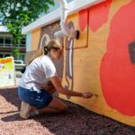 A woman kneels outside, painting a colorful mural with large flowers on a low wall during mural fest. A sign and building are visible in the background.