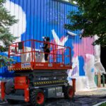 An artist on a lift paints a colorful mural on the side of a metal building during mural fest, with plastic sheets covering nearby surfaces and trees partially visible.