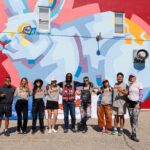 Ten people pose in front of a colorful geometric rabbit mural painted on a red wall, smiling and standing side by side in casual clothing at mural fest.
