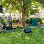 Two musicians perform with electric guitars and microphones under a tree in a grassy outdoor area during mural fest, with sound equipment and buildings visible in the background.