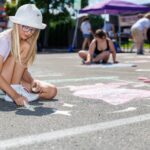 A young girl in a white hat and glasses draws with chalk on pavement at mural fest, with other people drawing in the background.