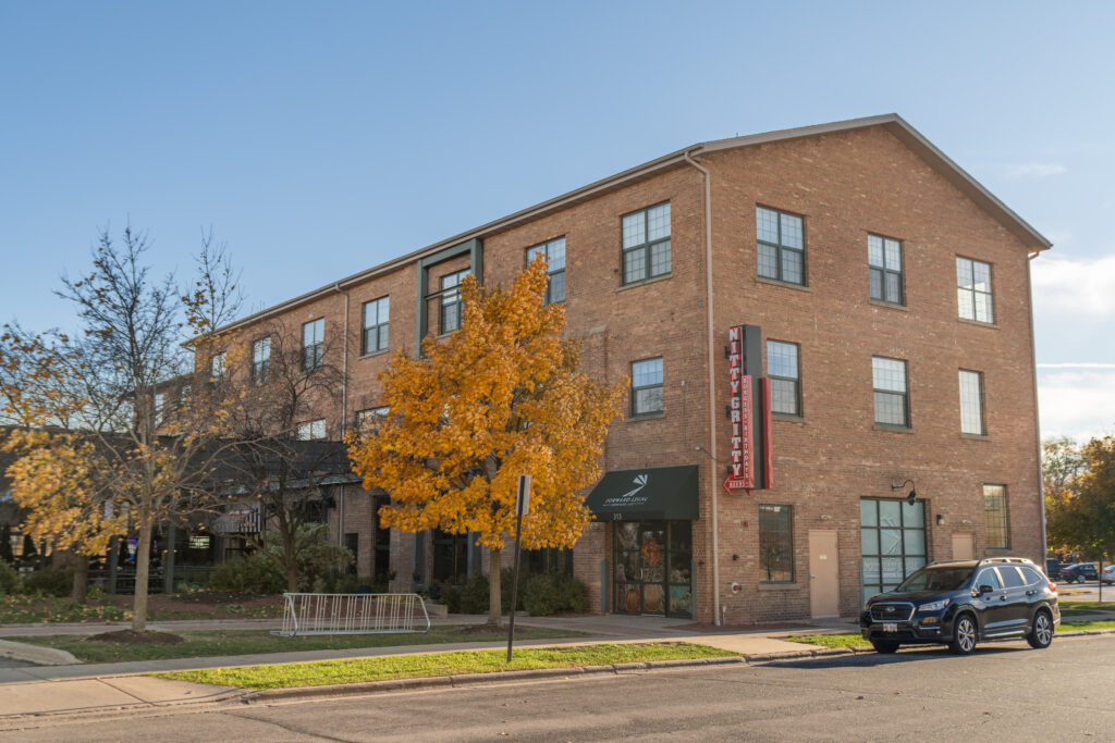 Three-story brick building with a Nitty Gritty restaurant sign, a tree with golden autumn leaves in front, and a parked SUV on the street.