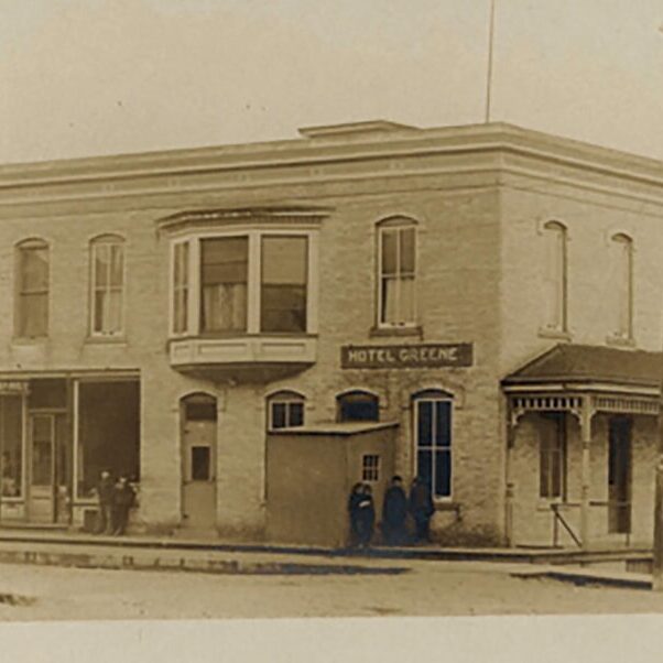 A sepia-toned photograph of Hotel Greene, an old two-story brick building with storefronts and a corner entrance. Four people stand outside; a horse and buggy are partially visible on the right.