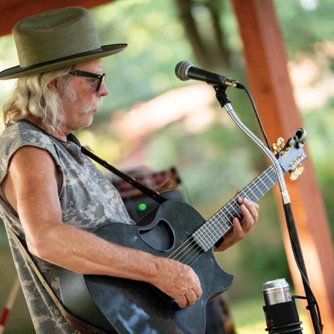 A person with long grey hair, wearing a hat and sunglasses, playing an acoustic guitar and standing in front of a microphone outdoors.