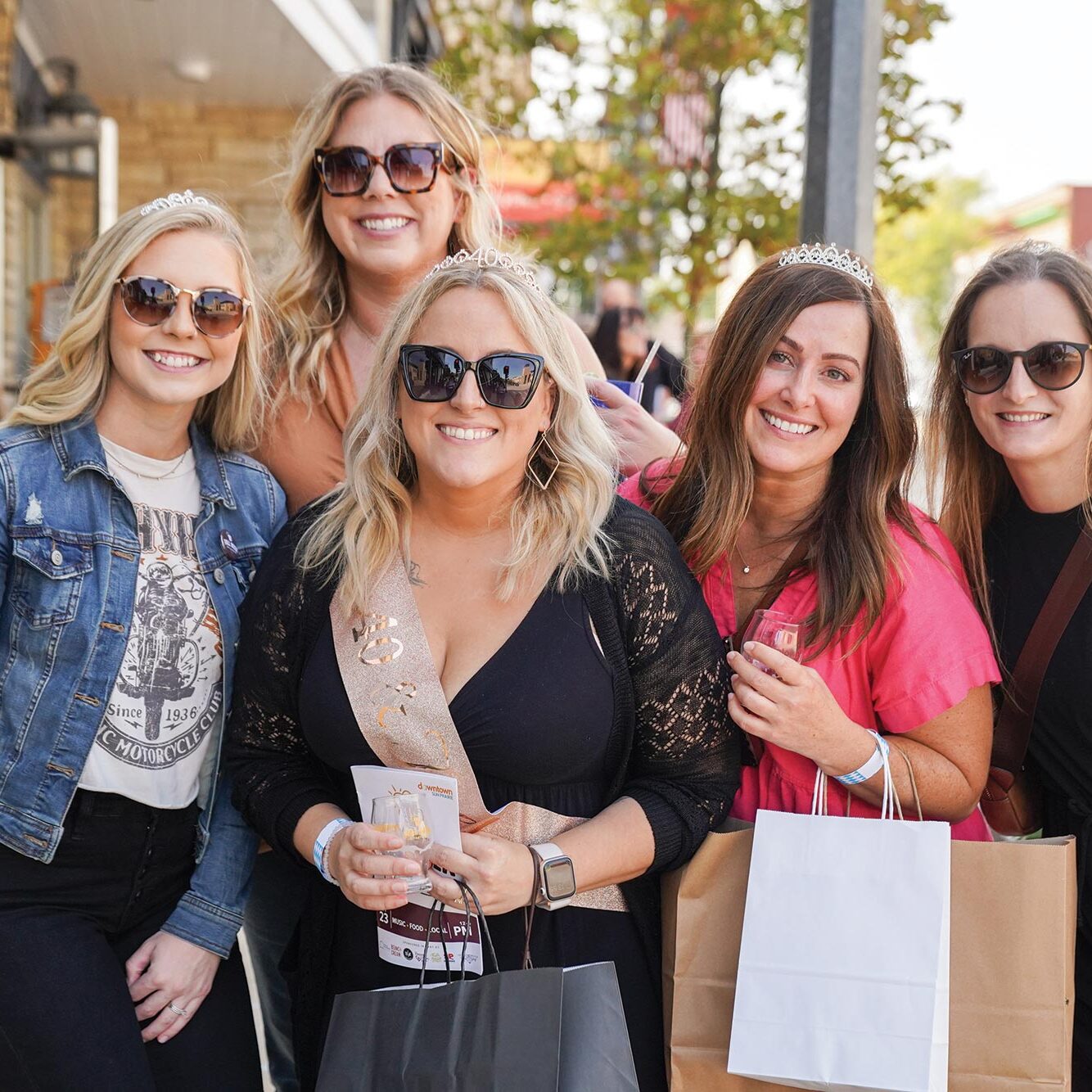 Five women wearing casual outfits and sunglasses stand close together outdoors, smiling at the camera. They are holding shopping bags and appear to be enjoying a sunny day.