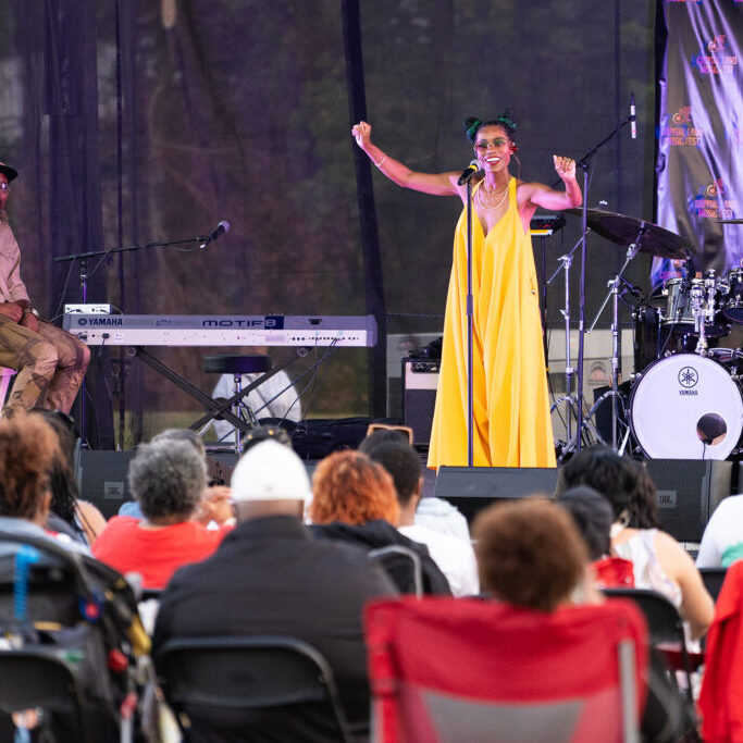 Outdoor concert with an audience seated on chairs, viewing a performer in a yellow dress singing on stage, surrounded by instruments and sound equipment, on a grassy field with trees in the background.