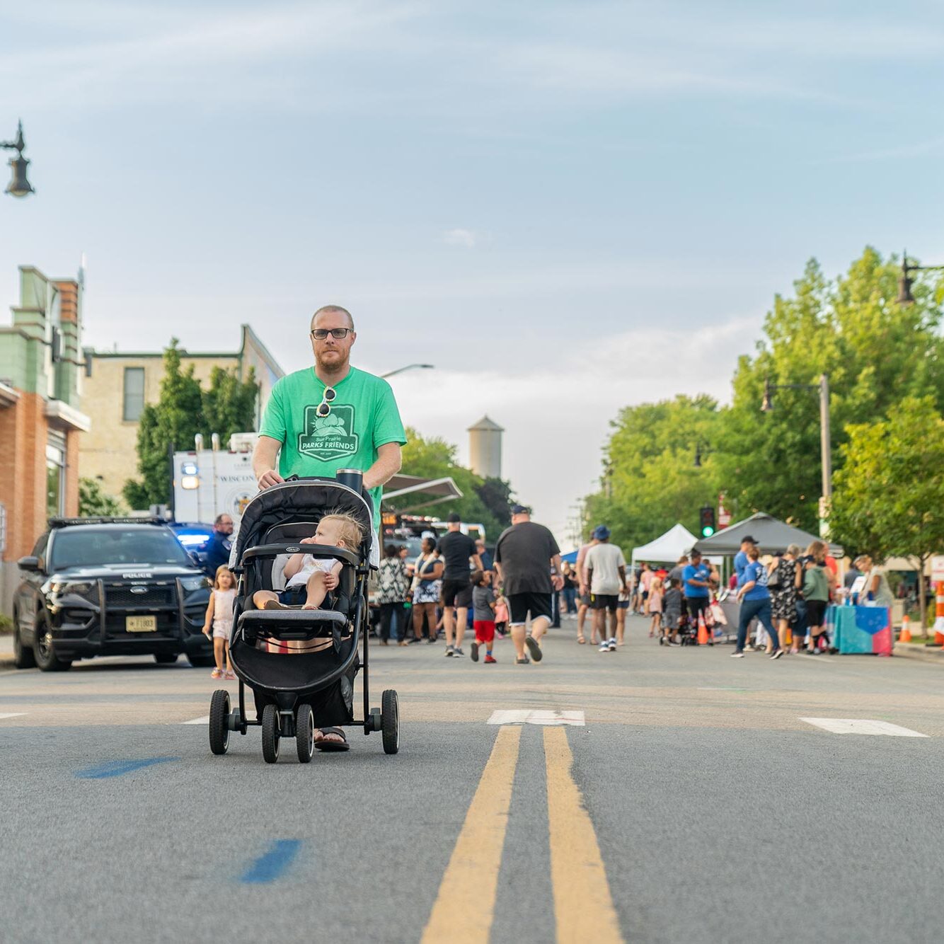 A man in a green shirt pushes a stroller down the middle of a street during a community event; people, booths, and a police car are visible in the background.