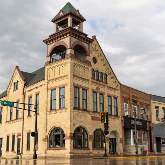 A historic brick building on a street corner under a cloudy sky, with traffic lights and wet pavement visible. The building features arched windows and a tower with a peaked roof.