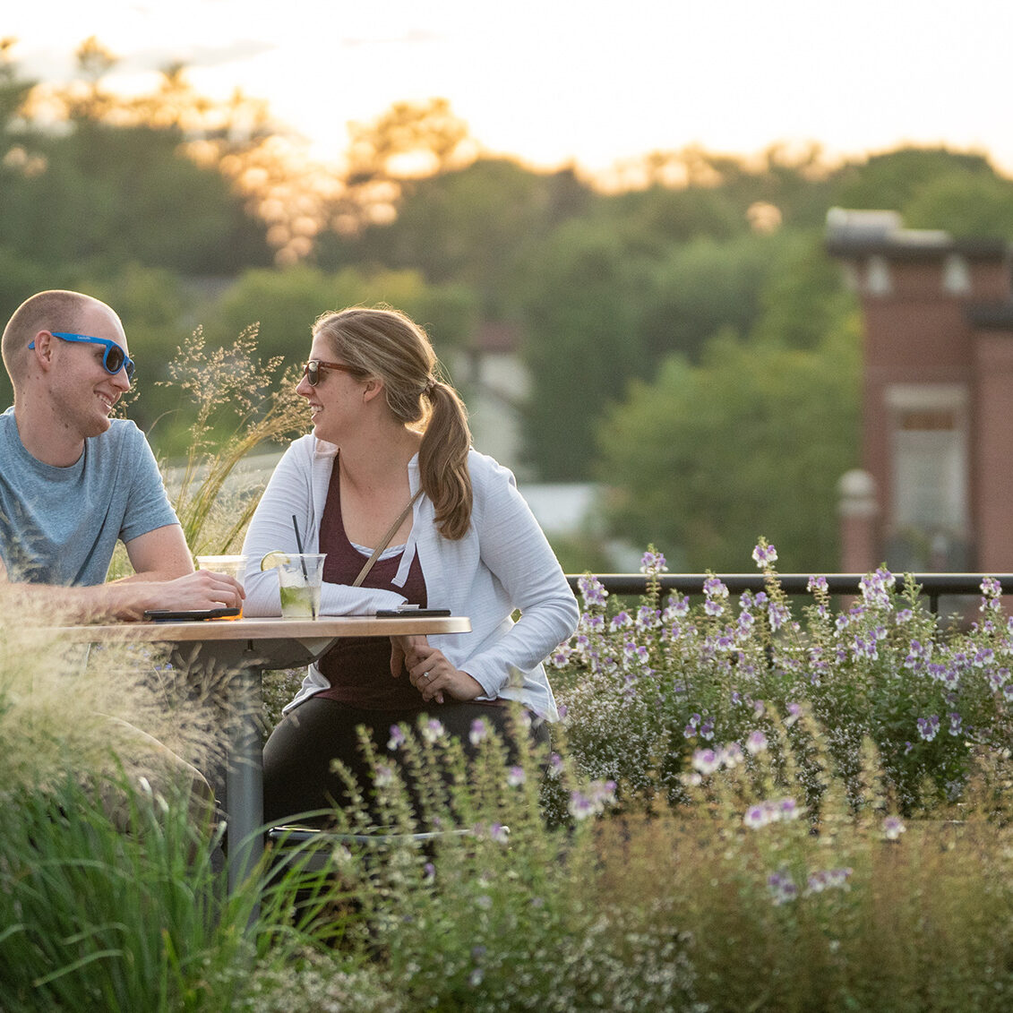 A man and a woman sit at a table on an outdoor patio surrounded by greenery, engaging in conversation during sunset.