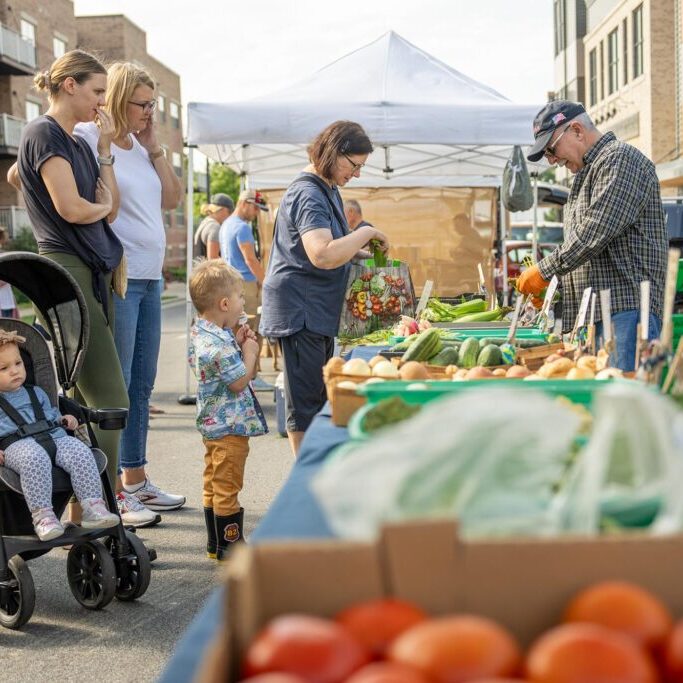 People browse fresh produce at an outdoor market. A woman pushes a stroller with a child, another woman stands nearby, and a man in a hat selects items from a stall.