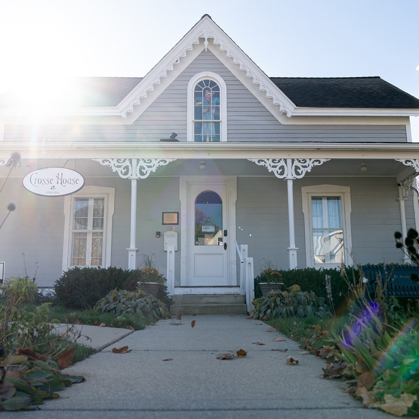 A quaint grey house with white trim, featuring a steep roof, decorative gables, a small porch, and a gated white picket fence, bathed in sunlight.