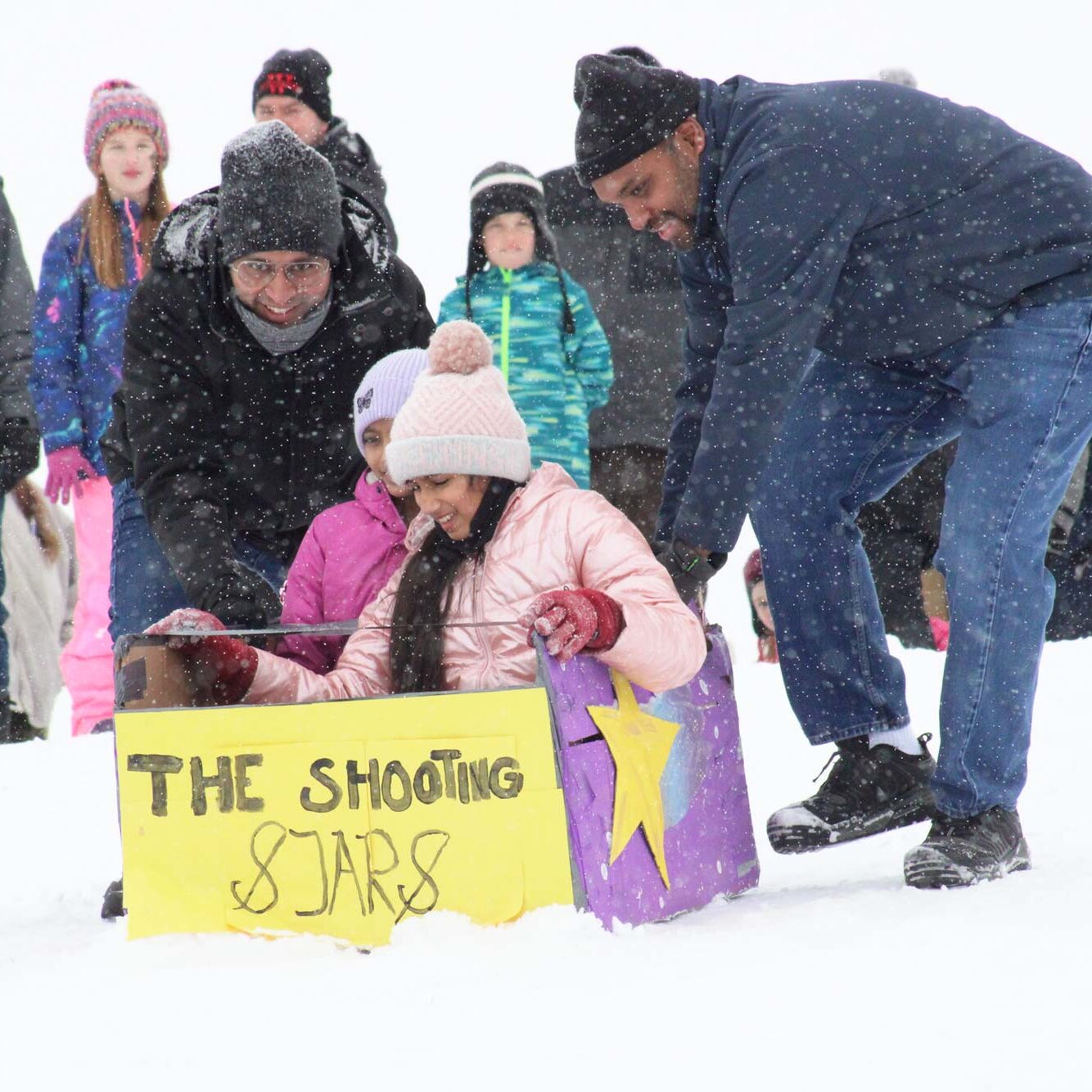 Two adults help two children in a cardboard sled labeled "The Shooting Stars" down a snowy hill, with a crowd of people in winter clothing watching in the background.