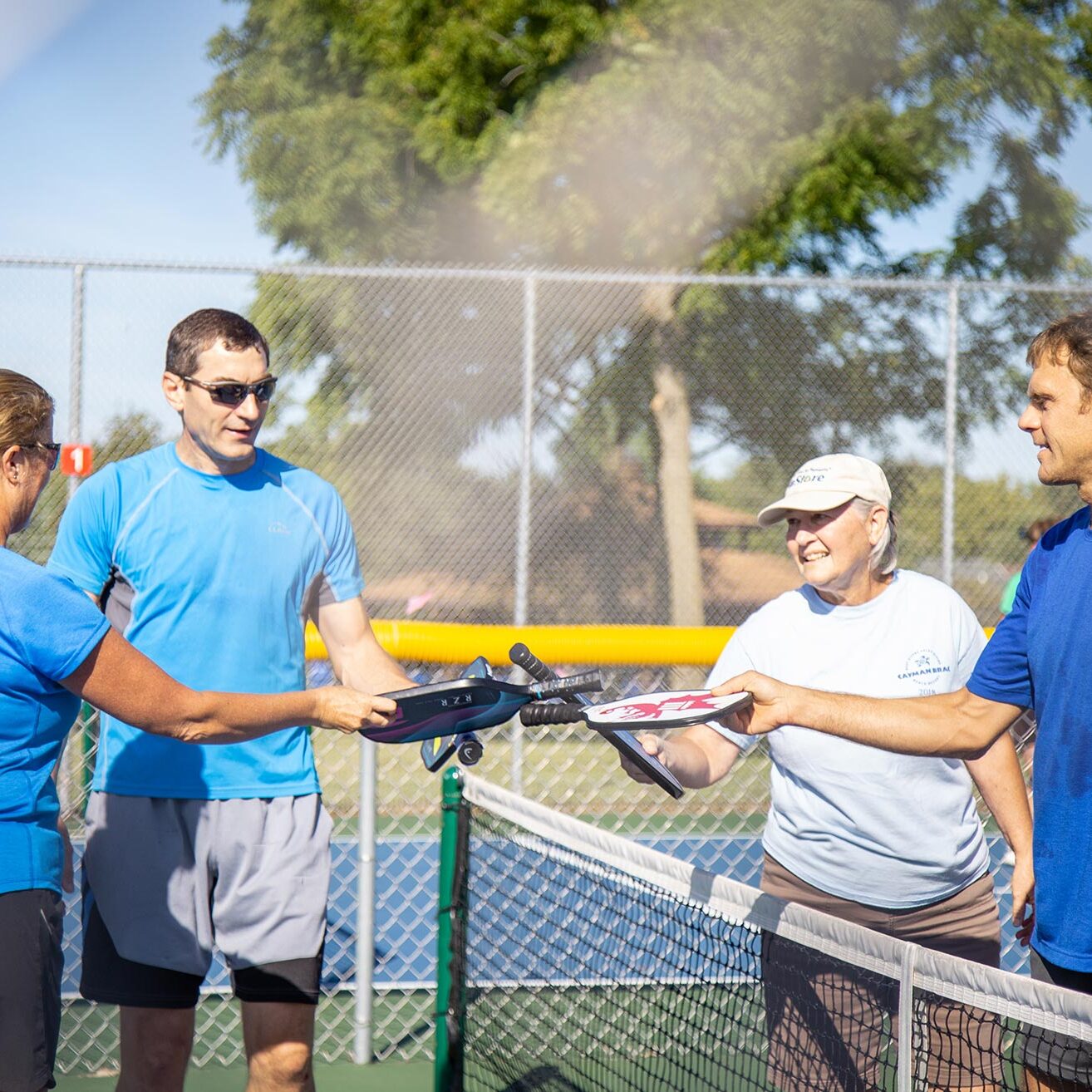 Four people on a tennis court shake hands over the net, wearing sports attire and holding rackets. They appear to have just finished a match.