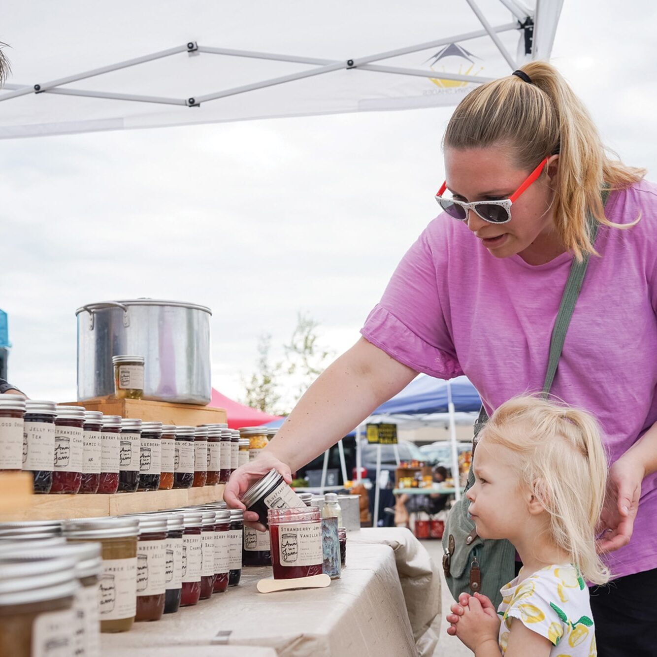 A woman in a pink shirt and red sunglasses selects a jar from a table of preserves, accompanied by a young child at an outdoor market. Another person stands behind the table near more jars.