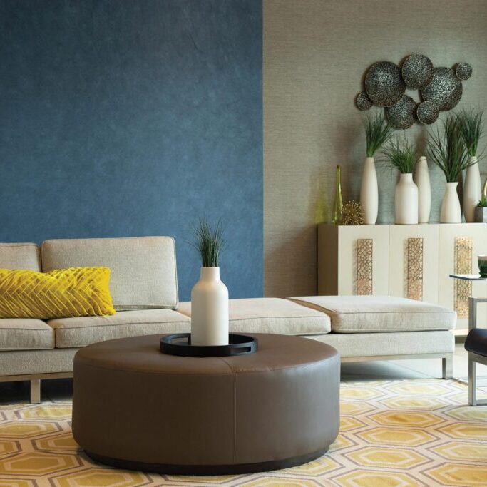 A modern living room featuring a beige sectional sofa with yellow and white pillows, a round brown ottoman, a white vase centerpiece, geometric wall art, and a blue accent wall.