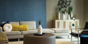 A modern living room featuring a beige sectional sofa with yellow and white pillows, a round brown ottoman, a white vase centerpiece, geometric wall art, and a blue accent wall.