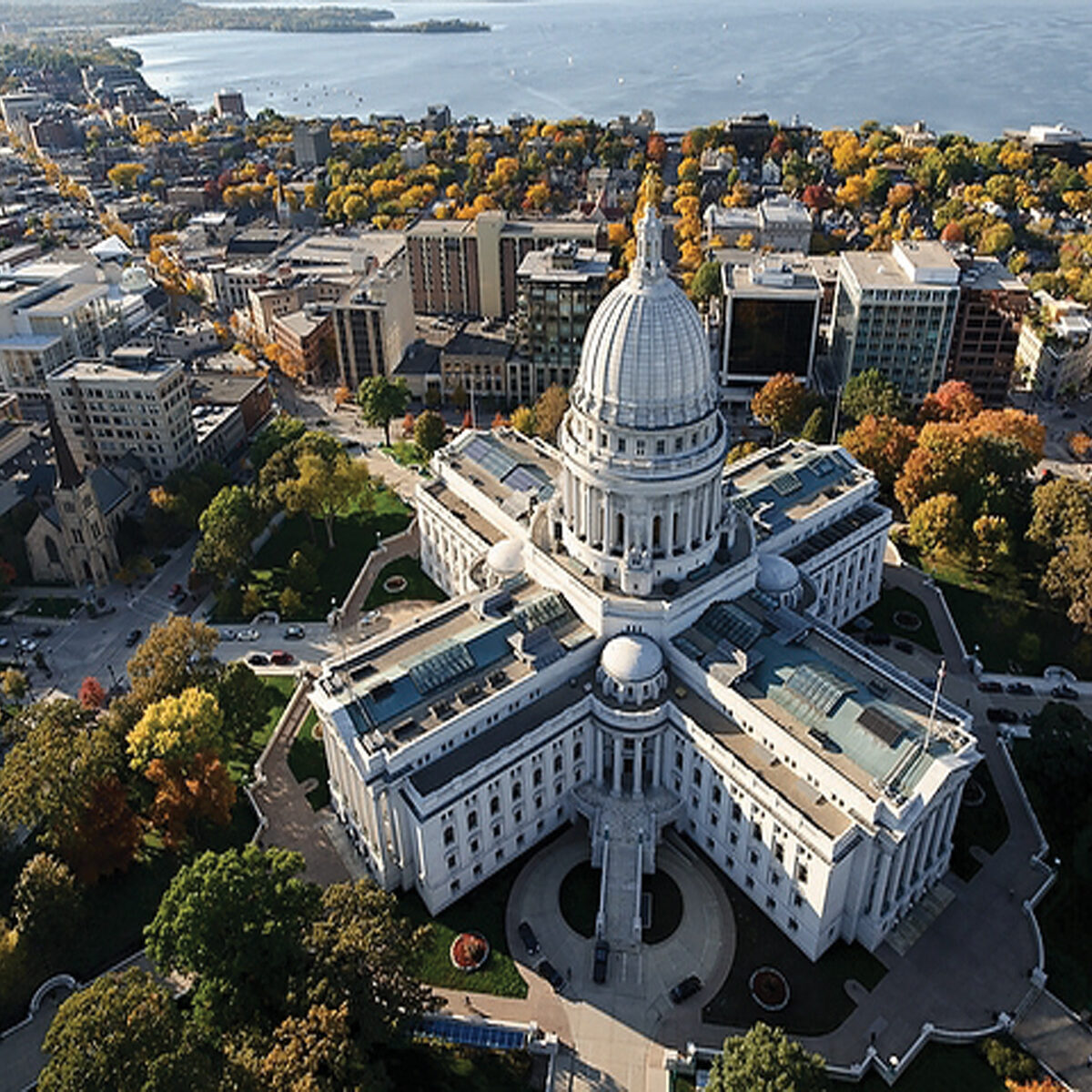 Aerial view of a domed capitol building surrounded by city streets, trees with autumn foliage, and a waterfront in the background.