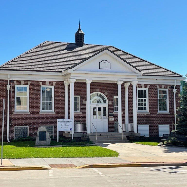 A red brick, single-story municipal building with white columns, a peaked roof, and American flags prominently displayed outside. The surrounding area includes a sidewalk, trees, and a streetlamp.