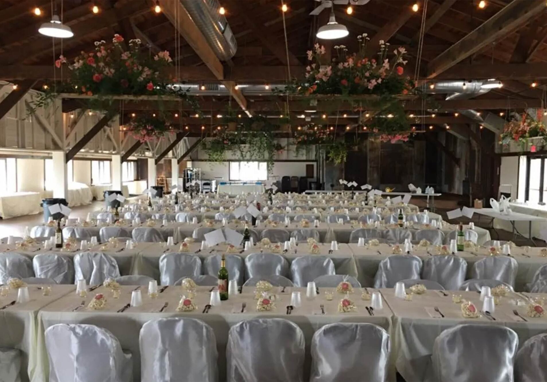 A large rustic hall is set up for an event with rows of tables and white chairs, floral centerpieces, and hanging flower decorations under a wooden ceiling with string lights.