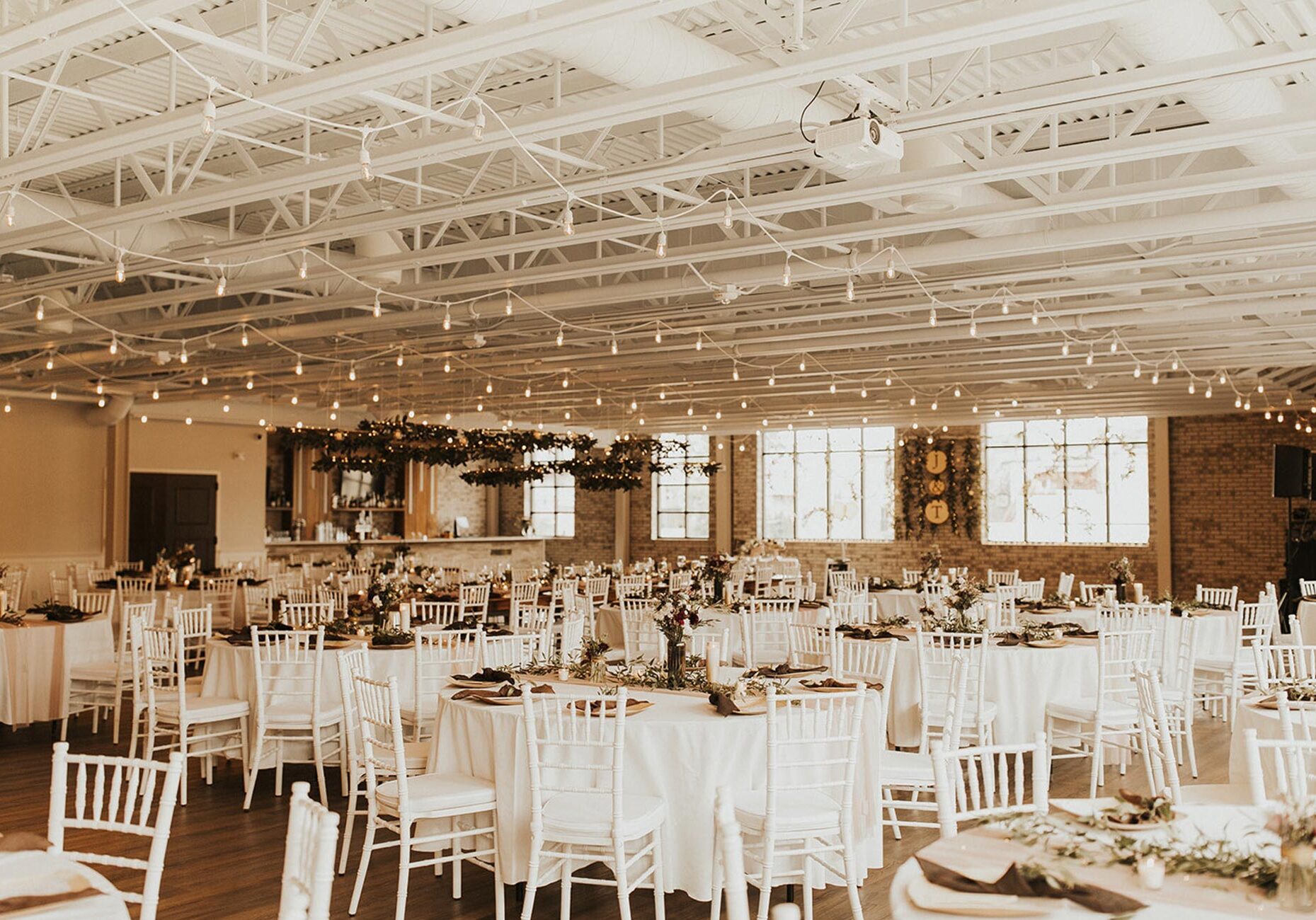 A spacious, rustic indoor event hall with white chairs and round tables elegantly set for a formal gathering. The ceiling is decorated with string lights, and natural light enters through large windows.