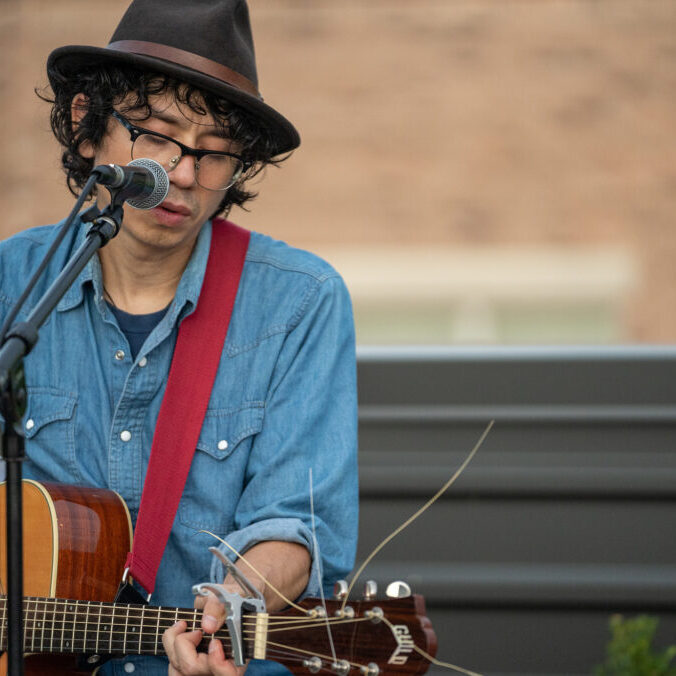 A person with curly hair, glasses, and a hat is playing an acoustic guitar and singing into a microphone. They are wearing a denim shirt and are outdoors in front of a blurred background.