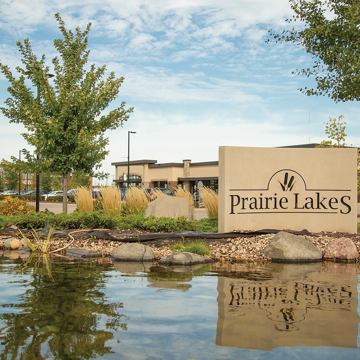 Outdoor sign for Prairie Lakes with trees and a pond in the foreground and buildings in the background.