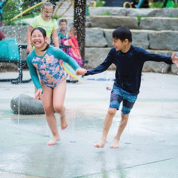 Two children in swimwear run through a splash pad, holding hands and laughing, with water spraying around them. Other children and adults can be seen in the background.